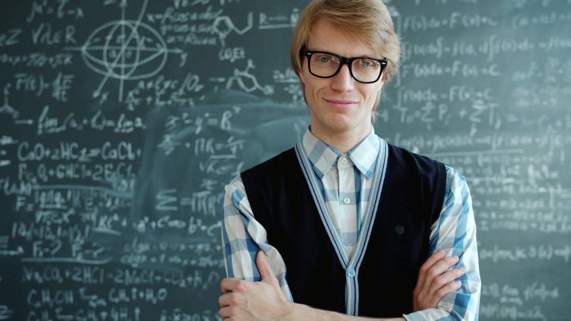 Un hombre rubio con gafas sonríe con los brazos cruzados delante de una pizarra escolar completamente saturada de fórmulas científicas.
